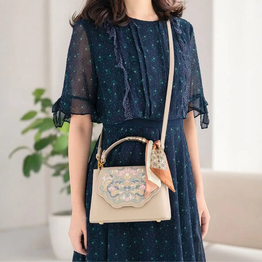East Asian woman carries beige new Chinese embroidered crossbody handbag with silk scarf, wearing navy floral chiffon dress in bright minimalist indoor room