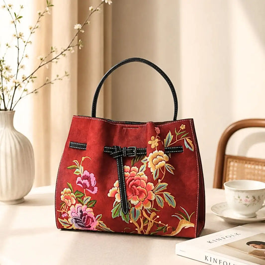Retro red leather tote with peony floral embroidery placed on a beige table beside a teacup, book and flowering vase in soft natural light.