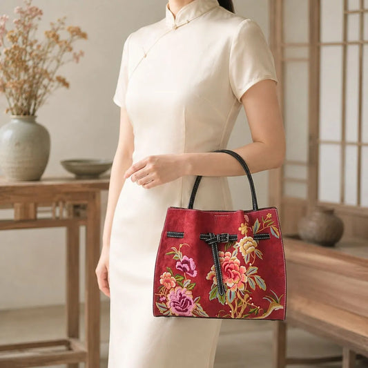 Red peony embroidered leather handbag held by a woman wearing beige cheongsam in a traditional Chinese wooden interior room.