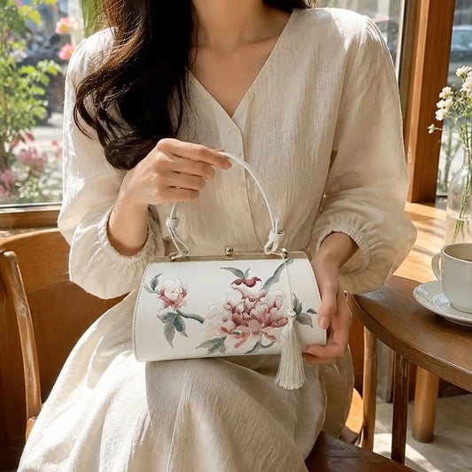 White barrel handbag with peony embroidery and tassel, held by a lady in beige linen dress at bright window cafe for casual oriental outfit inspiration.