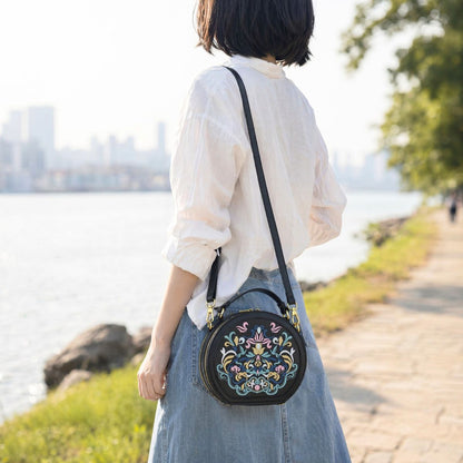 Back shot of Asian woman in white linen shirt and denim long skirt, wearing black national style embroidered round bag beside riverside walkway, showing adjustable shoulder strap and side bag shape