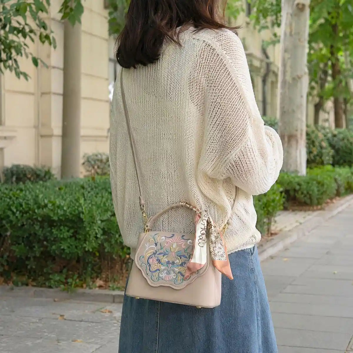 Back view of woman in loose knit top and denim skirt, showing shoulder wearing beige national style embroidered handbag on city sidewalk