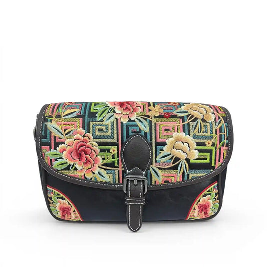 Black PU leather flap crossbody bag featuring retro geometric pattern and vibrant multi-tonal peony floral embroidery, finished with black leather buckle detail against a clean white studio backdrop.
