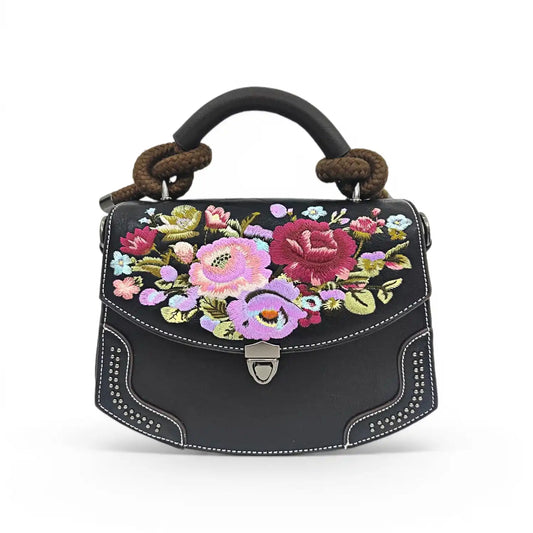 Black PU leather flap handbag decorated with colorful vintage wildflower embroidery, rivet trim and braided rope top handle, presented on a clean plain white background with retro metal turn-lock hardware.