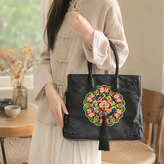 Black ethnic embroidered top handle tote held by lady in beige traditional linen blouse in bright cozy indoor room for daily casual oriental styling.