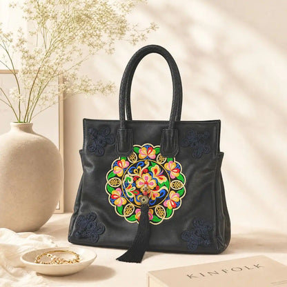 Black leather tote bag with colorful circular ethnic embroidery and tassel, placed on warm indoor table with vase and book to show craft details.