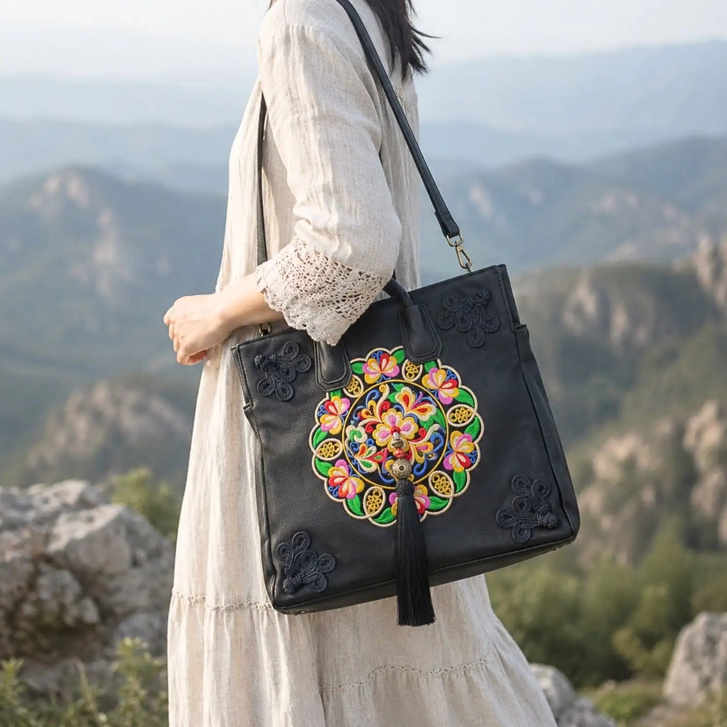 Black leather ethnic embroidered tote worn crossbody with detachable shoulder strap by woman in linen dress against mountain landscape for travel lifestyle.