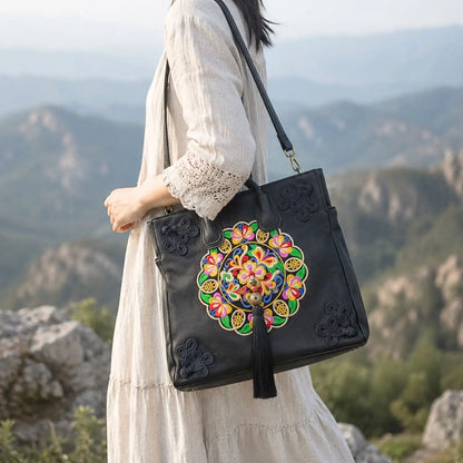 Black leather ethnic embroidered tote worn crossbody with detachable shoulder strap by woman in linen dress against mountain landscape for travel lifestyle.