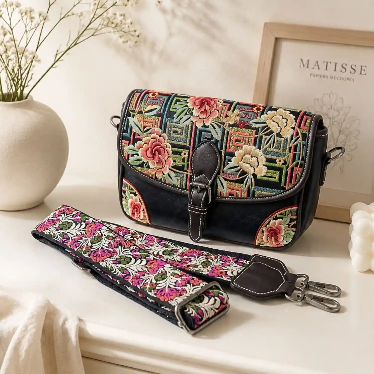 Black peony geometric embroidered saddle crossbody bag displayed on bright tabletop, shown with its detachable adjustable floral wide shoulder strap and home decor.