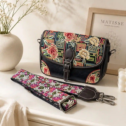 Black peony geometric embroidered saddle crossbody bag displayed on bright tabletop, shown with its detachable adjustable floral wide shoulder strap and home decor.