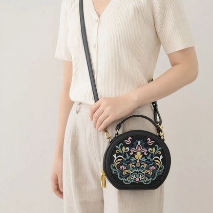 East Asian woman wears cream ribbed knit top and linen trousers, crossbody black round leather handbag with traditional colorful floral embroidery and golden zipper hardware, minimalist light beige indoor background, casual commuter lifestyle shot