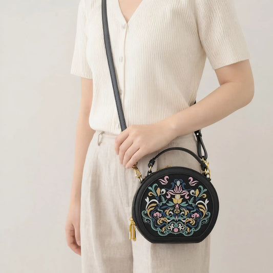East Asian woman wears cream ribbed knit top and linen trousers, crossbody black round leather handbag with traditional colorful floral embroidery and golden zipper hardware, minimalist light beige indoor background, casual commuter lifestyle shot