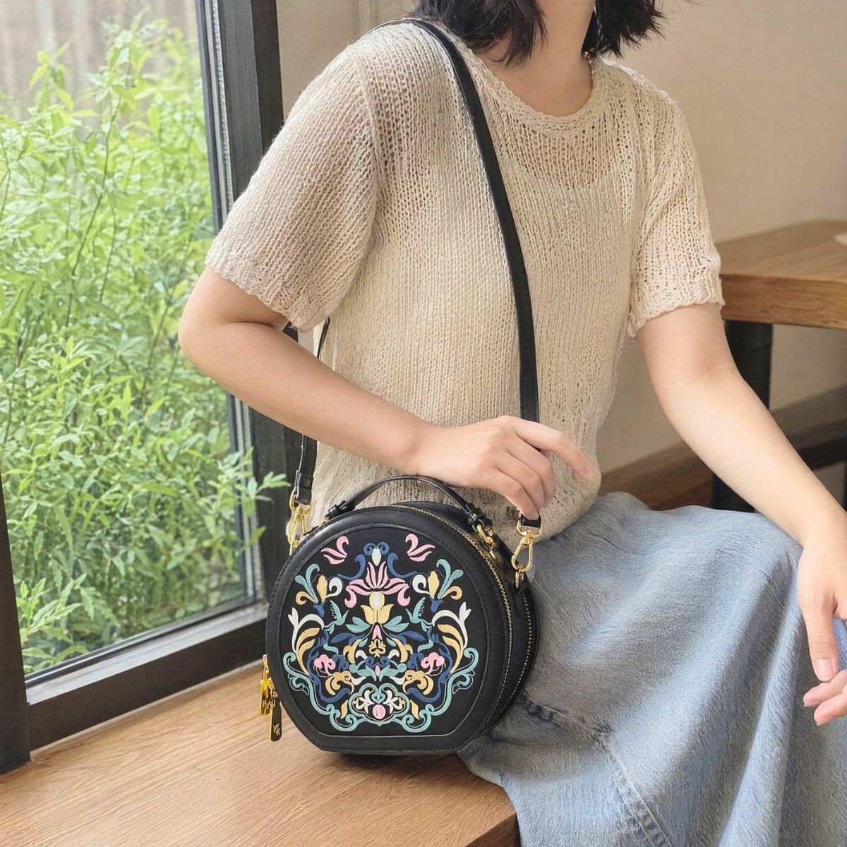 Woman in hollow beige knit top and denim midi skirt sits by green plant window, crossbody vintage Chinese embroidered round leather bag, warm soft indoor leisure lifestyle photo