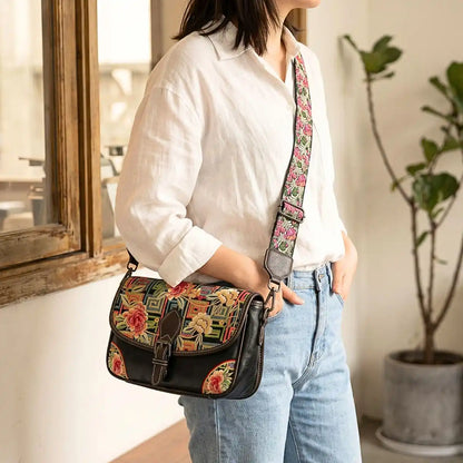 Ethnic peony embroidered black saddle crossbody crossbody bag worn with white linen shirt and blue jeans beside wooden window, ideal for casual everyday street fashion. 