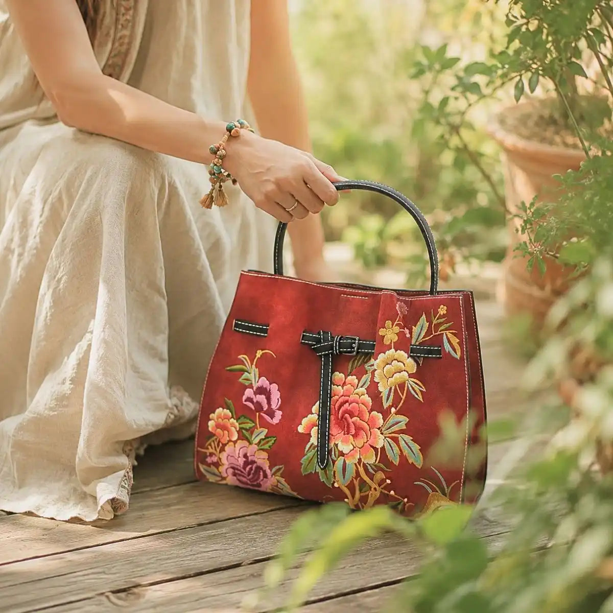 Red peony embroidered leather handbag held by a lady in loose linen dress on wooden deck surrounded by lush green garden plants.