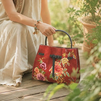 Red peony embroidered leather handbag held by a lady in loose linen dress on wooden deck surrounded by lush green garden plants.
