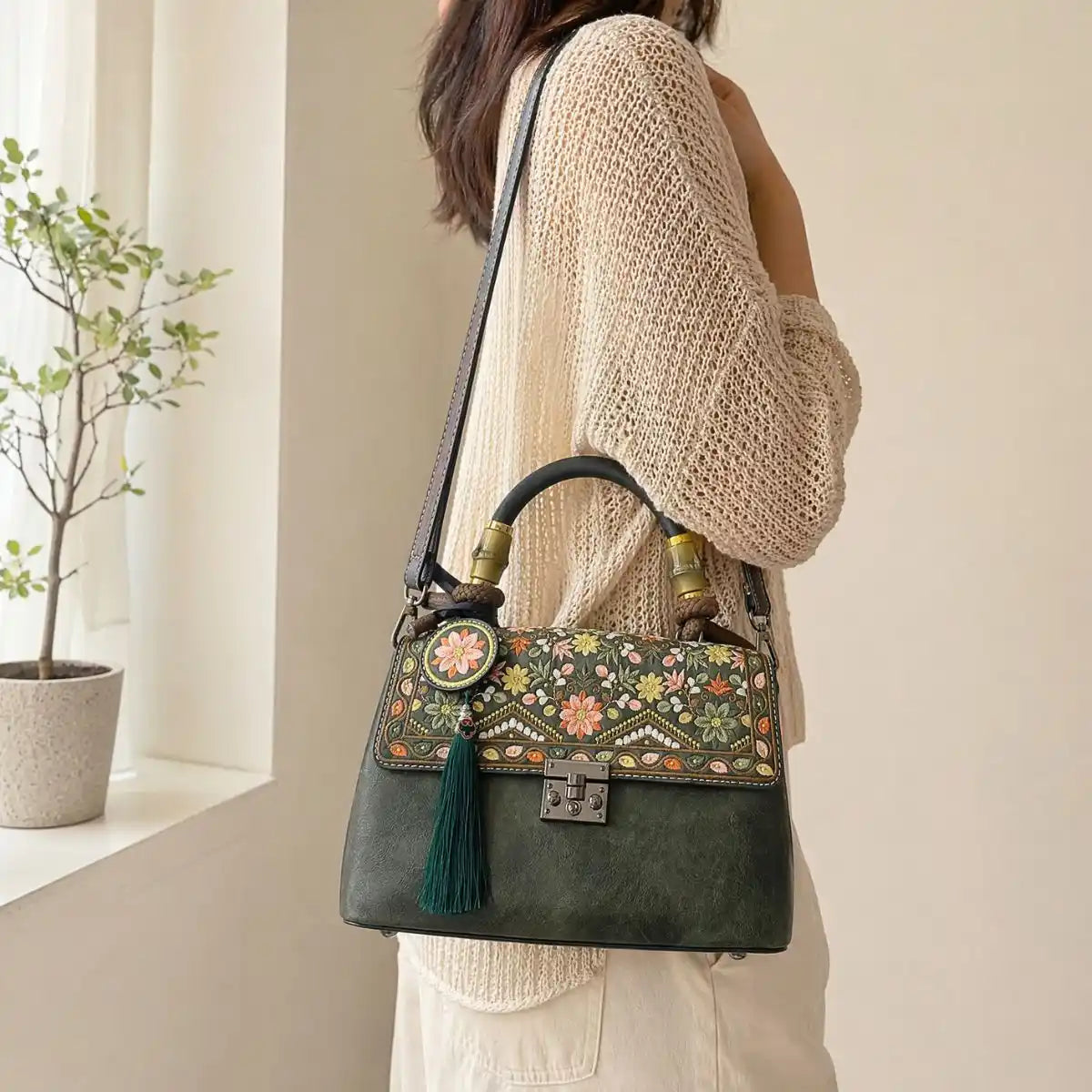 Faceless Asian woman wears beige hollow knit cardigan, crossbody dark green vintage floral handbag with bamboo handle and green tassel, warm minimalist indoor window background