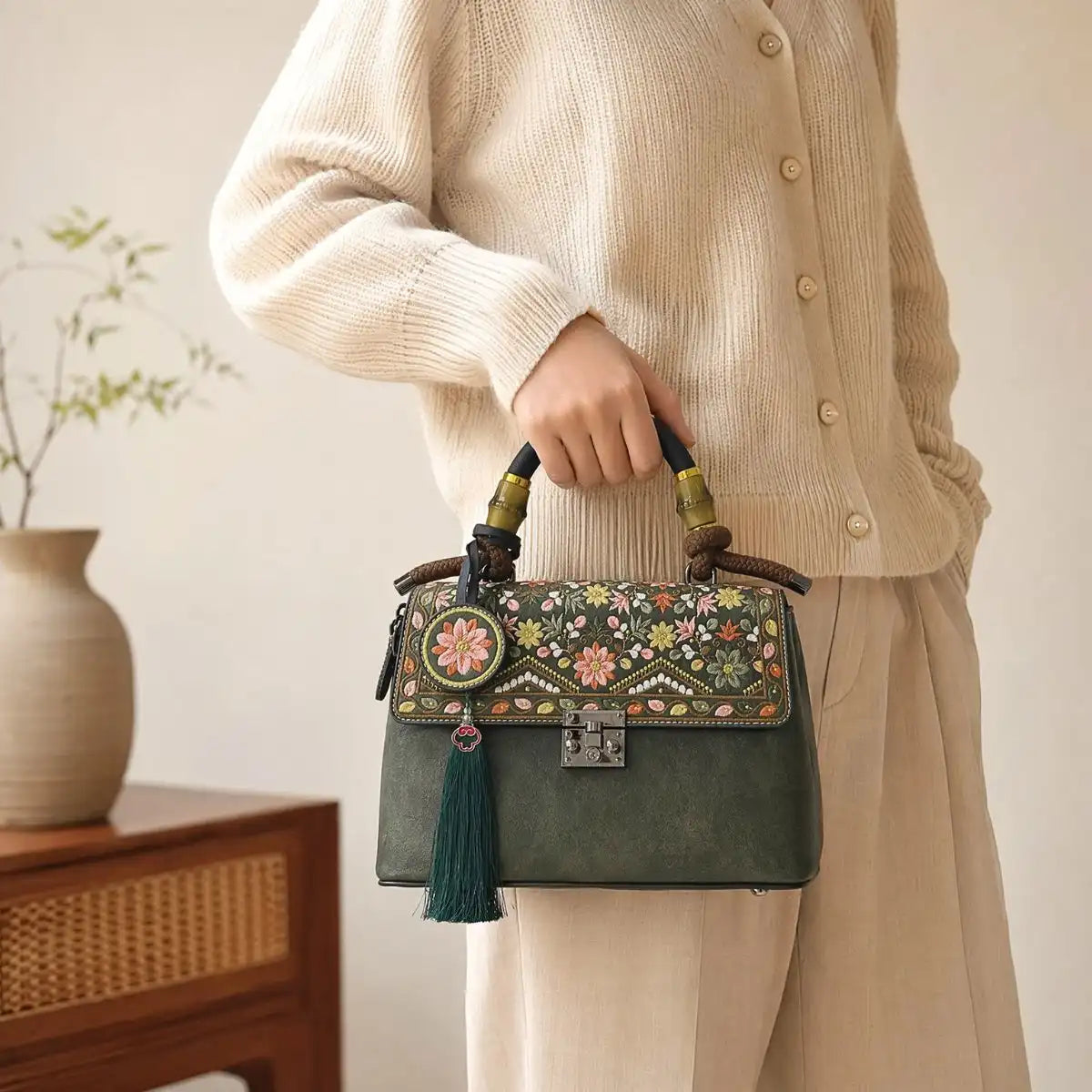 Faceless Asian woman holds bamboo handle of dark green embroidered handbag, wearing cream knit cardigan, simple wooden home interior with ceramic vase decor, soft natural light