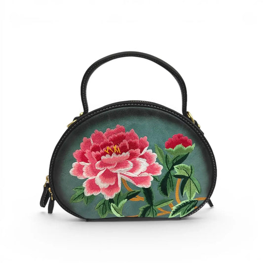 Dark green round PU leather top handle handbag decorated with intricate vibrant pink peony floral embroidery and green leaves, shot on a clean plain white studio background with polished gold hardware details.