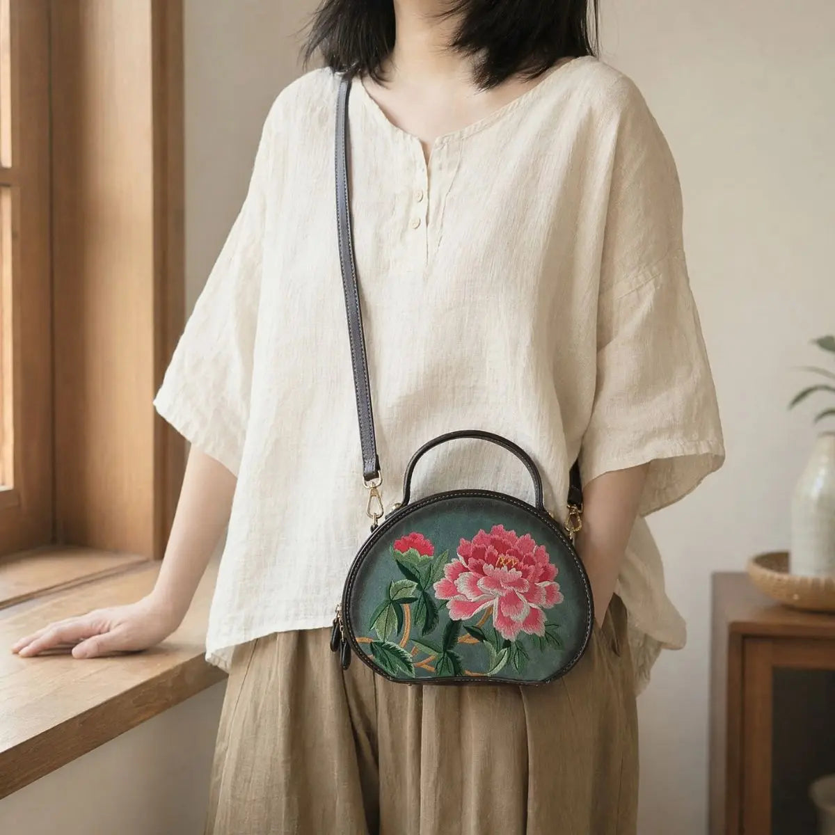 Woman wearing loose beige short-sleeve linen top and wide-leg casual trousers, styled with dark green round PU leather crossbody bag featuring vivid pink peony floral embroidery by a wooden window.