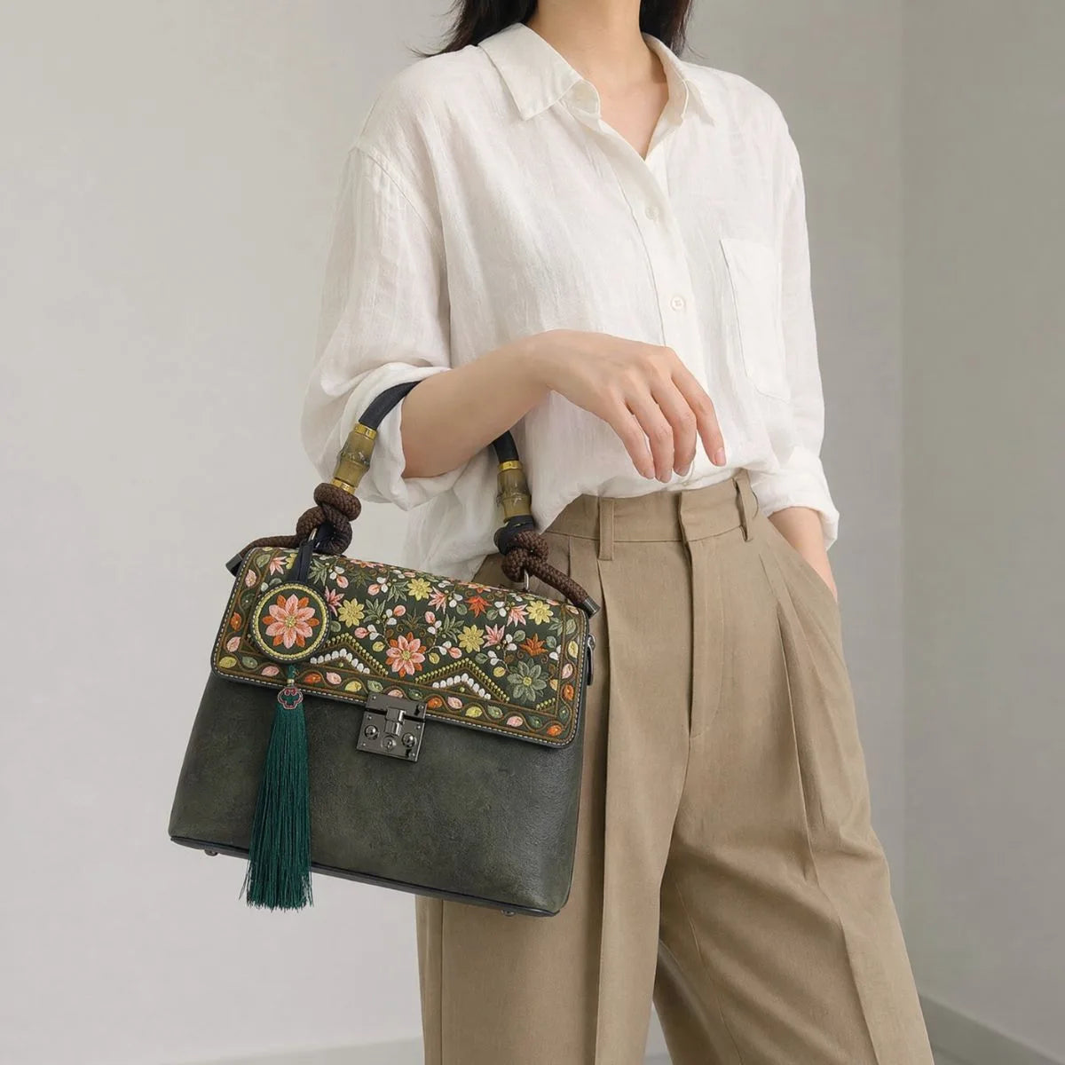 Faceless Asian woman carries dark green vintage handbag under arm, wearing white linen shirt and khaki trousers, plain light gray indoor workplace background
