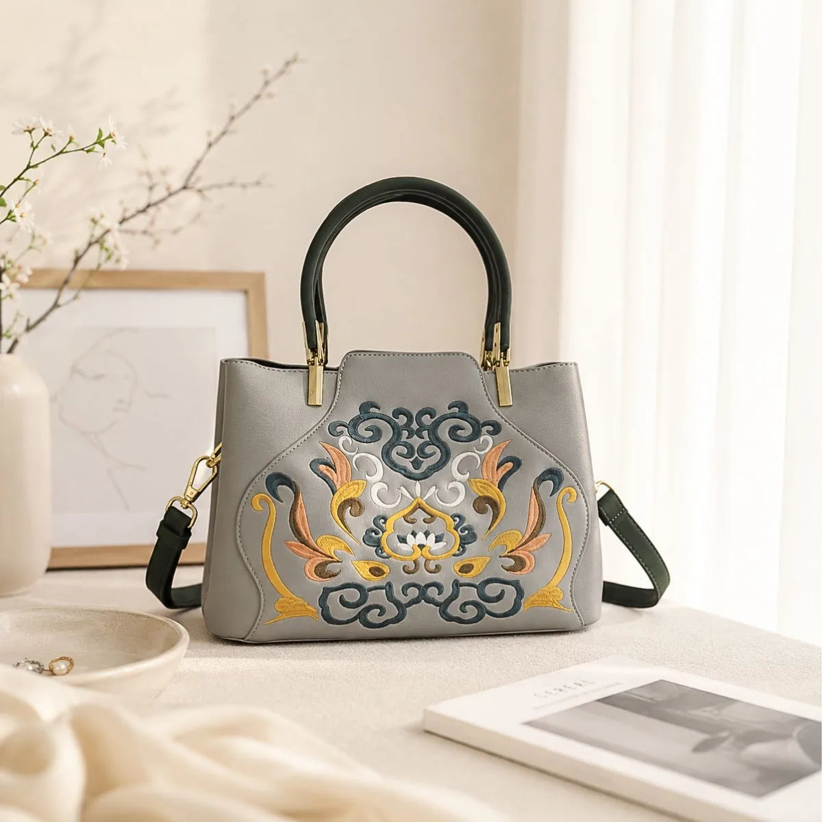 Gray leather handbag with traditional scroll floral embroidery placed on warm beige table beside flower branches, book and jewelry for product detail display.
