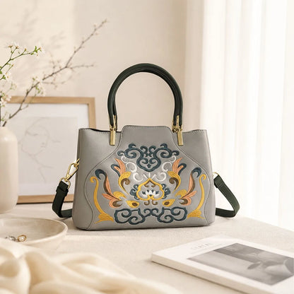 Gray leather handbag with traditional scroll floral embroidery placed on warm beige table beside flower branches, book and jewelry for product detail display.