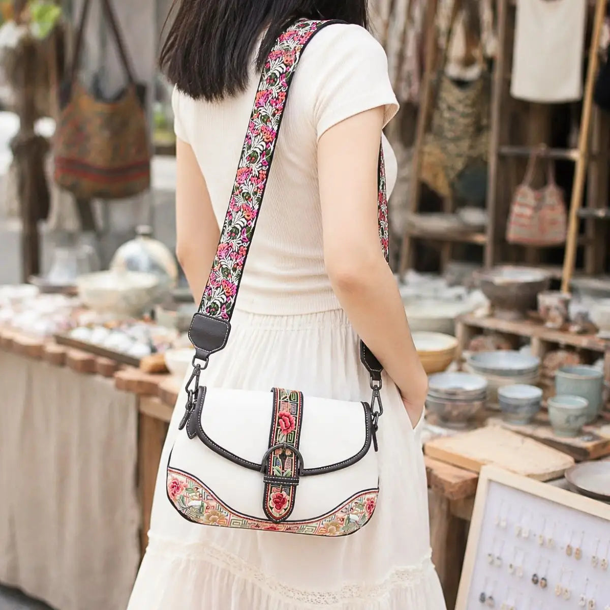 White boho saddle crossbody bag with vibrant floral embroidery, carried by lady in casual white dress at traditional handicraft market for travel outfit inspiration.