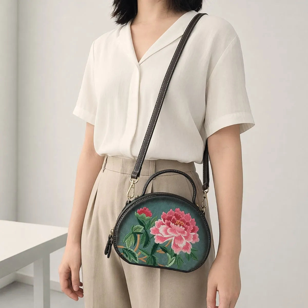 Woman in white lapel linen short sleeve shirt and high-waisted khaki trousers, carrying dark green round PU leather handbag decorated with delicate pink peony embroidery in minimalist indoor space.