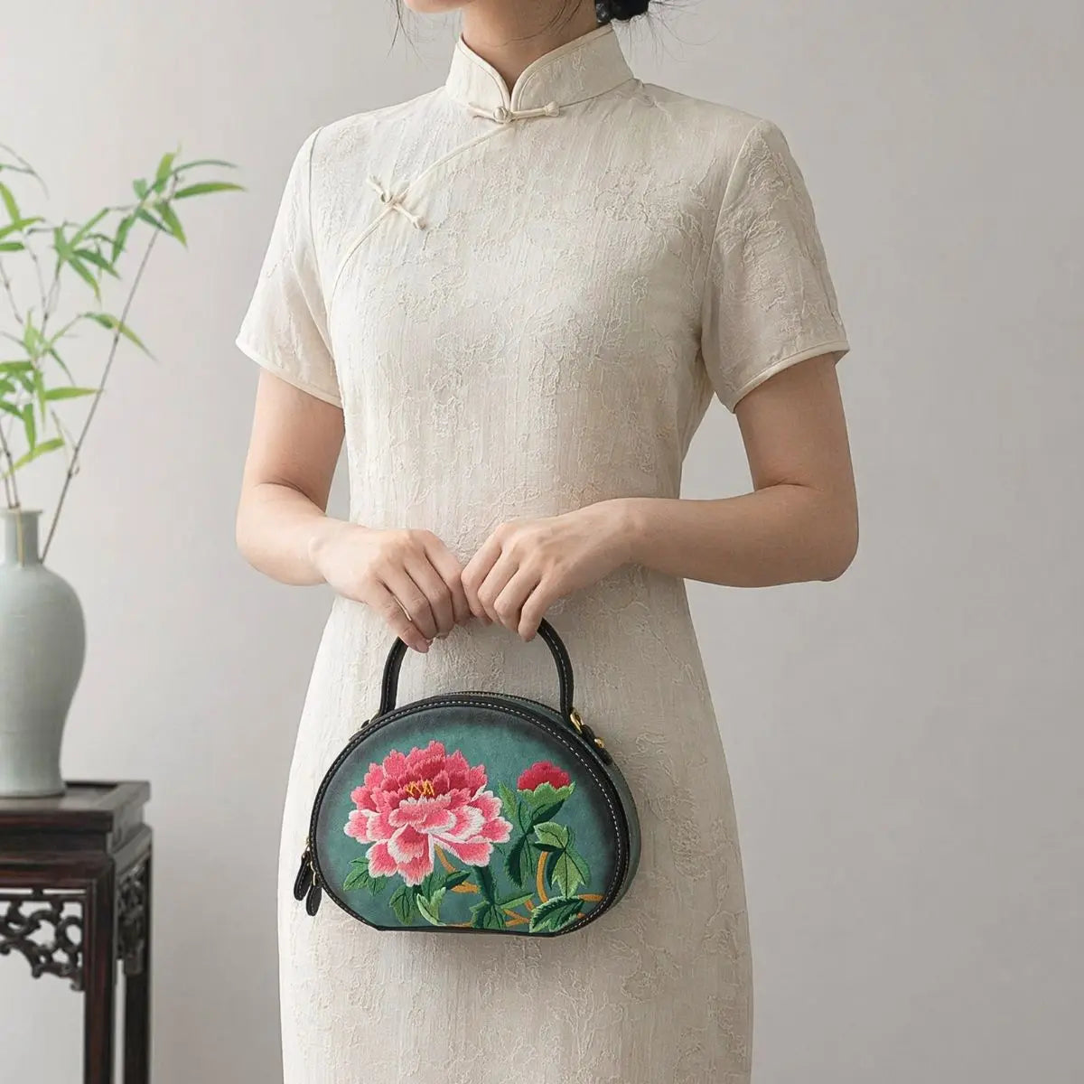 Woman dressed in cream textured traditional linen cheongsam, holding a dark green round top handle PU leather bag with elaborate pink peony floral embroidery beside bamboo decor.