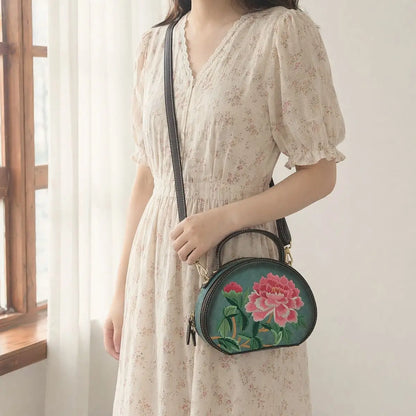 Lady wearing cream vintage puff sleeve floral midi dress, accessorized with dark green round PU leather crossbody bag stitched with bright pink peony blooms near sheer window curtains.