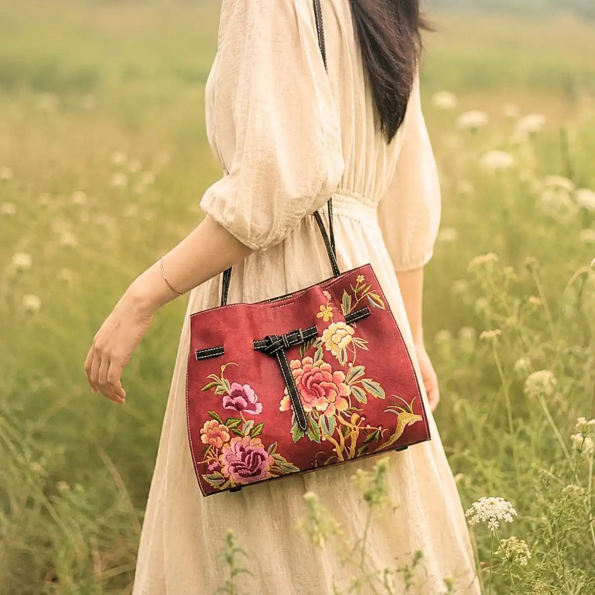 Red leather tote bag with colorful peony embroidery carried by a lady in beige linen dress, posing in a wild grass field for oriental outfit inspiration.