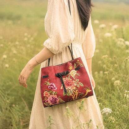 Red leather tote bag with colorful peony embroidery carried by a lady in beige linen dress, posing in a wild grass field for oriental outfit inspiration.