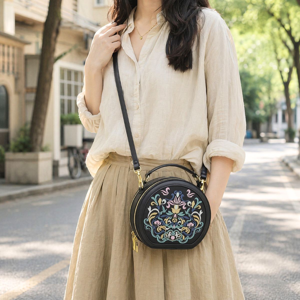 Woman in beige linen shirt and khaki long skirt carries black embroidered round crossbody bag on tree-lined city street, casual literary daily outfit, clear bag embroidery and golden hardware details