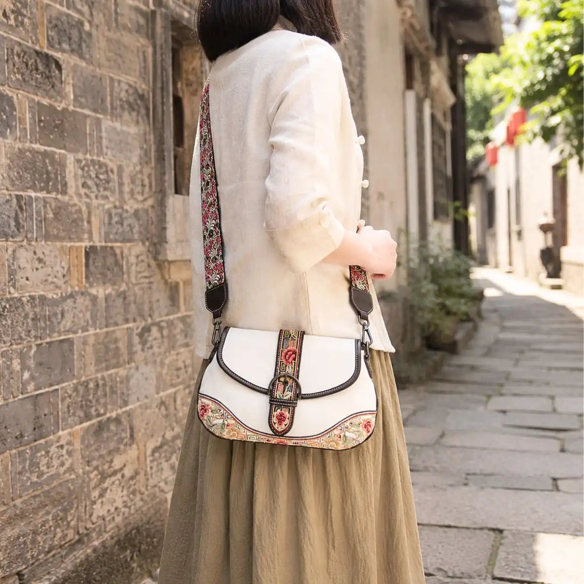 White ethnic embroidered crossbody saddle bag matched with beige linen top and long skirt, shot in old stone alley to show retro oriental casual style.