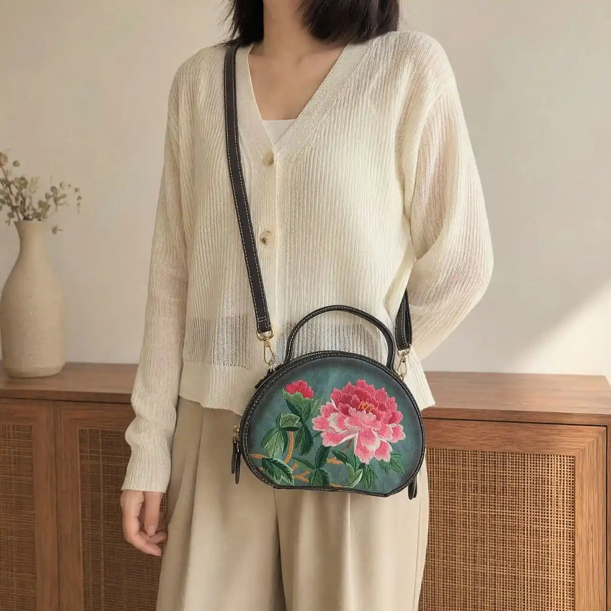 Woman wearing cream ribbed V-neck knit cardigan and khaki wide-leg trousers, paired with dark green round PU leather crossbody bag with detailed pink peony floral embroidery in warm neutral home interior.