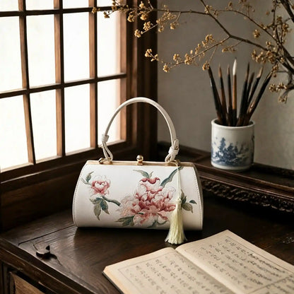 White peony embroidered barrel handbag placed on wooden desk beside calligraphy brushes and ancient books by traditional wooden lattice window.
