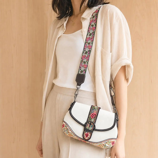 White vintage saddle crossbody bag with colorful ethnic floral embroidery and wide embroidered shoulder strap, worn with casual linen outfit for daily boho styling.