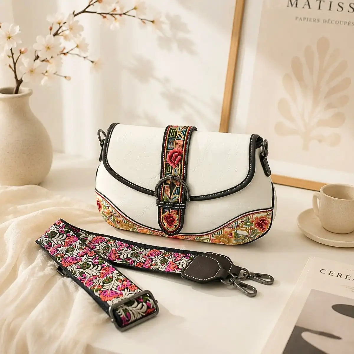 White vintage saddle crossbody bag with detachable floral embroidered wide strap, displayed on bright indoor table to showcase bag details and accessory.