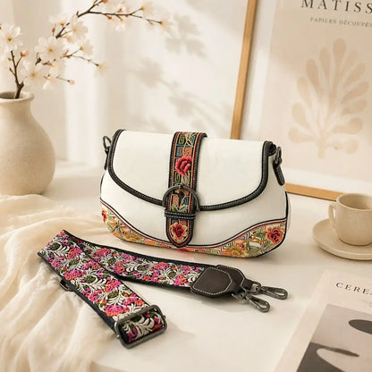 White vintage saddle crossbody bag with detachable floral embroidered wide strap, displayed on bright indoor table to showcase bag details and accessory.